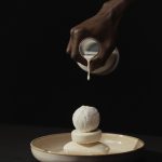 Main d'une personne à la peau foncée versant de la crème sur une pile de savons en barre sur une assiette en céramique dans un cadre minimaliste.