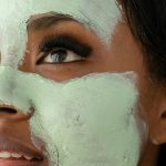 Crop d'une femme afro-américaine méconnaissable avec un masque d'argile verte sur le visage, regardant vers le haut pendant une routine quotidienne de soins de la peau dans une pièce lumineuse