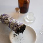 Une nature morte représentant un bâton de sauge en train de brûler sur une soucoupe accompagnée de bouteilles en verre vintage.