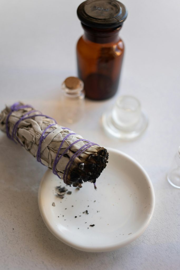 Une nature morte représentant un bâton de sauge en train de brûler sur une soucoupe accompagnée de bouteilles en verre vintage.