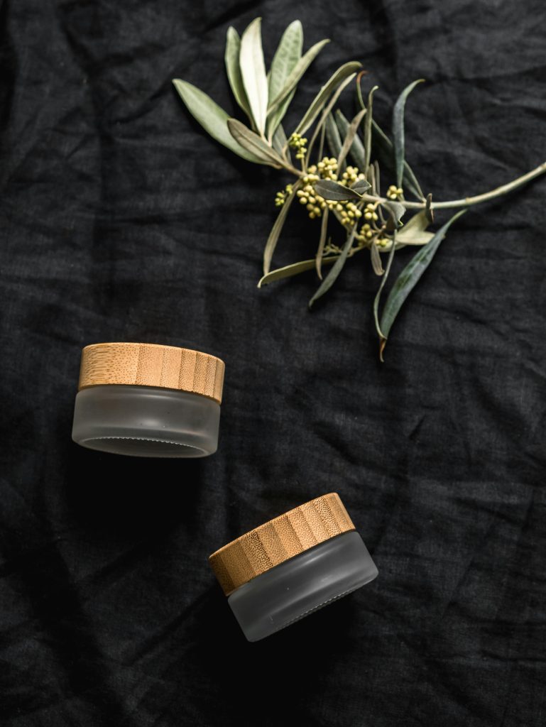 Organic cosmetic jars with wooden lids on black fabric and olive branches, vue de dessus