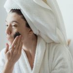 Une femme applique un masque facial dans le cadre de sa routine matinale de soins de la peau, enveloppée dans une serviette douce.
