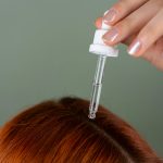Gros plan de l'application de sérum sur les racines avec un compte-gouttes de précision sur des cheveux roux, illustrant une routine de soin capillaire.