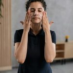 A woman in casual attire applying facial cream indoors as part of a skincare routine. Une femme en tenue décontractée appliquant une crème pour le visage à l'intérieur dans le cadre d'une routine de soins de la peau.
