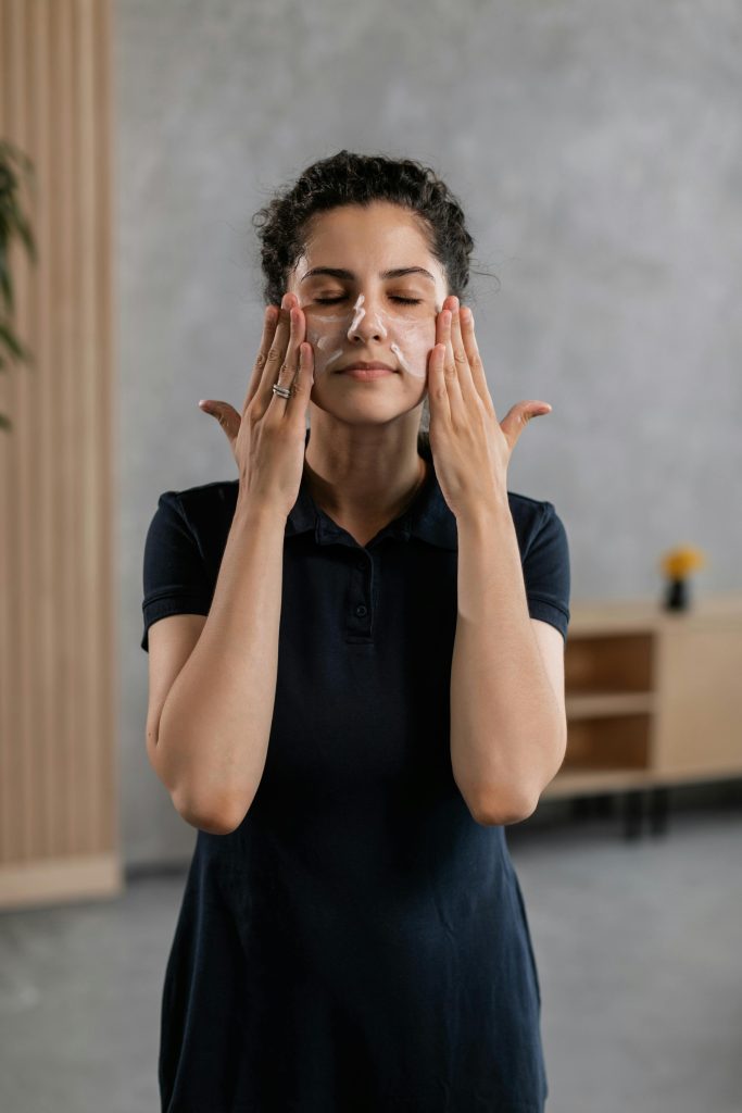 A woman in casual attire applying facial cream indoors as part of a skincare routine. Une femme en tenue décontractée appliquant une crème pour le visage à l'intérieur dans le cadre d'une routine de soins de la peau.