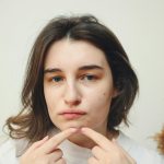 A young woman with acne examining a pimple on her cheek in a thoughtful expression. Une jeune femme atteinte d'acné examinant un bouton sur sa joue avec une expression réfléchie.