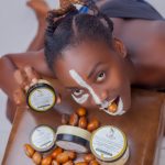 African woman biting nut, posing with natural shea skincare products and cream jars. Femme africaine mordant une noix, posant avec des produits de soin à base de karité naturel et des pots de crème.