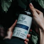 Hands holding a shampoo bottle against a leafy background, emphasizing natural hair care. Des mains tenant une bouteille de shampooing sur un fond feuillu, mettant l'accent sur les soins naturels des cheveux.