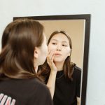 Young woman examines her skin in a mirror for a daily beauty routine. Jeune femme examinant sa peau dans un miroir pour sa routine beauté quotidienne.
