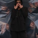 Individual in dark attire surrounded by transparent plastic and reaching hands, creating suspenseful atmosphere. Personne vêtue de noir entourée de plastique transparent et de mains qui tendent vers elle, créant une atmosphère de suspense.